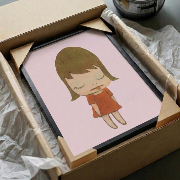 May include: Framed art print featuring a cute cartoon-style girl with brown bob haircut and closed eyes, wearing an orange dress, set against a soft pink background. The print is displayed in a black frame with cardboard packaging materials and wooden frame corners visible around it on a white surface.