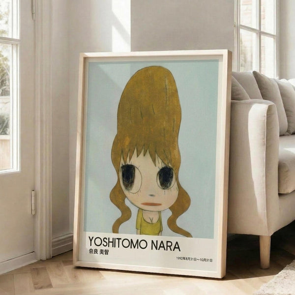 May include: Framed poster print featuring anime-style illustration of young girl with large golden blonde hair in ponytail, oversized dark eyes, and yellow top against soft blue background. Text reads "YOSHITOMO NARA" with Japanese characters and dates "1992年8月31日〜10月31日". Light wood frame leans against white wall next to beige sectional sofa in bright, minimalist living room with large windows.
