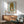 May include: Framed poster of cartoon-style girl with large brown hair buns, pale skin, and oversized eyes on mint green background with "YOSHITOMO NARA" text below. Wooden frame hangs above mid-century modern walnut credenza with white ceramic lamp. Sage green upholstered armchair with wooden arms sits beside small round table. Warm sunlight streams across white walls creating geometric shadows.