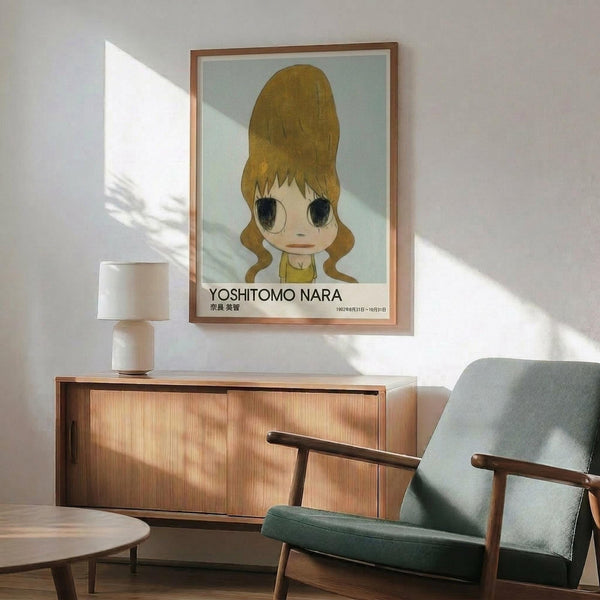 May include: Framed poster of cartoon-style girl with large brown hair buns, pale skin, and oversized eyes on mint green background with "YOSHITOMO NARA" text below. Wooden frame hangs above mid-century modern walnut credenza with white ceramic lamp. Sage green upholstered armchair with wooden arms sits beside small round table. Warm sunlight streams across white walls creating geometric shadows.