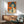 May include: A framed vintage Japanese Coca-Cola advertisement poster featuring a woman with dark bob haircut holding a Coke bottle against an orange background with blue Japanese text. The poster hangs above a mid-century modern wooden credenza with a white table lamp, next to a teal upholstered armchair with wooden arms in a sunlit room with white walls.