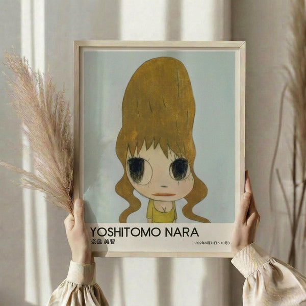 May include: Person in cream sweater holding framed art print featuring anime-style girl with blonde hair in yellow top, styled in minimalist exhibition poster format with artist name and dates below. Dried pampas grass visible on left side against neutral wall background.