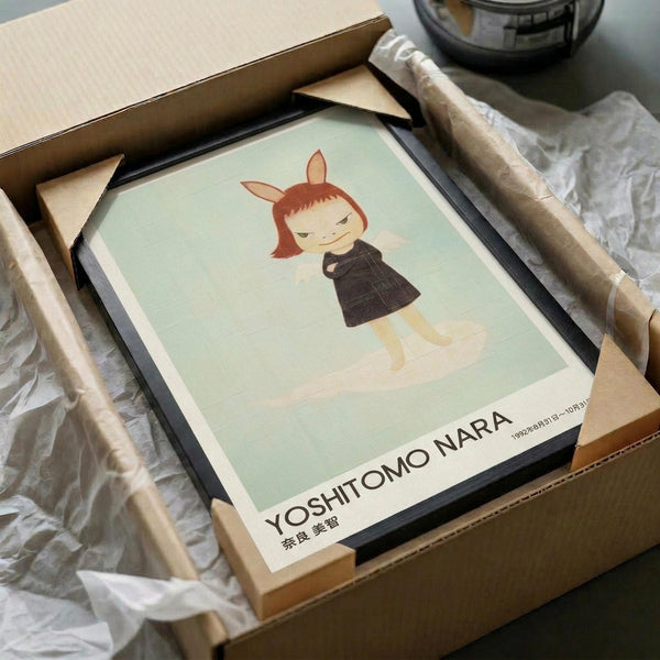 May include: Framed art print featuring a child-like figure with reddish hair and small horns wearing a dark dress against a pale green background. The print is displayed in a black frame with white matting, partially unpacked from brown cardboard packaging with white tissue paper. Wooden frame corners and packing materials are visible around the artwork.