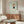 May include: Framed poster displayed above wooden sideboard in modern living room with white curved chair and glass coffee table. Artwork shows cartoon-style girl character with bunny ears, red bob haircut, and black dress on mint green background. Exhibition poster format with artist name at bottom. Warm wood tones throughout furniture pieces, soft natural lighting, minimalist Scandinavian-inspired interior design.