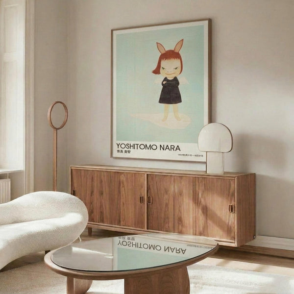 May include: Framed poster displayed above wooden sideboard in modern living room with white curved chair and glass coffee table. Artwork shows cartoon-style girl character with bunny ears, red bob haircut, and black dress on mint green background. Exhibition poster format with artist name at bottom. Warm wood tones throughout furniture pieces, soft natural lighting, minimalist Scandinavian-inspired interior design.