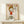 May include: Framed poster featuring a cartoon-style character with blonde bob haircut wearing red dress, making an angry expression with raised middle finger. Bold red letters spell out profanity above the figure with small yellow star. Artist name "Yoshitomo Nara" appears below in black text with Japanese characters and exhibition dates. Light wood frame sits on pale wooden shelf next to small white wrapped gift with twine and greenery. Minimalist white wall background with soft natural lighti