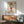May include: Framed poster featuring cartoon character with blonde hair in red dress making middle finger gesture, with profanity text above and artist name below. Displayed on white wall above wooden sideboard with table lamp, next to mid-century modern green upholstered chair with wooden arms. Bright natural lighting creates geometric shadows across the minimalist interior scene.