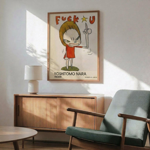 May include: Framed poster featuring cartoon character with blonde hair in red dress making middle finger gesture, with profanity text above and artist name below. Displayed on white wall above wooden sideboard with table lamp, next to mid-century modern green upholstered chair with wooden arms. Bright natural lighting creates geometric shadows across the minimalist interior scene.
