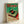 May include: Framed vintage Japanese advertisement poster featuring a turntable record player with green and red color scheme, displayed against white wall. The retro-style artwork shows detailed turntable illustration with Japanese text, housed in light wooden frame, leaning at slight angle on floor or surface.