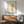 May include: Framed artwork showing a cartoon-style child with blonde bob haircut and green eyes wearing a light blue sleeveless outfit, displayed above a wooden sideboard with table lamp in a mid-century modern living room setting with teal upholstered chair and natural lighting creating shadows on white walls.