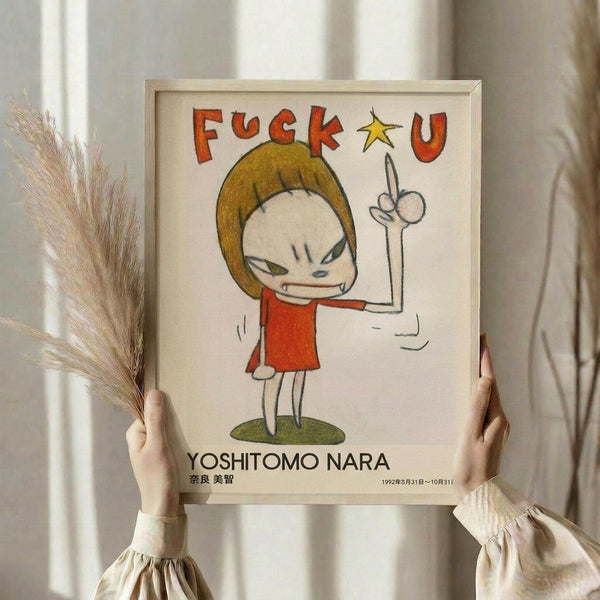May include: Hands holding a framed poster featuring a cartoon-style angry child character with blonde bob haircut in red dress, giving middle finger gesture. Text reads "FUCK U" in red letters above figure, with yellow star accent. Artist name "YOSHITOMO NARA" appears below artwork in black text with Japanese characters and dates. Light wood frame against neutral beige background with dried pampas grass visible on left side.