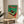 May include: Framed vintage-style poster with Japanese text and illustrated turntable in green and red color scheme, displayed on round wooden side table next to beige upholstered chair. Natural lighting creates soft shadows on white wall. Plant leaves visible in corner, creating modern minimalist room setting.