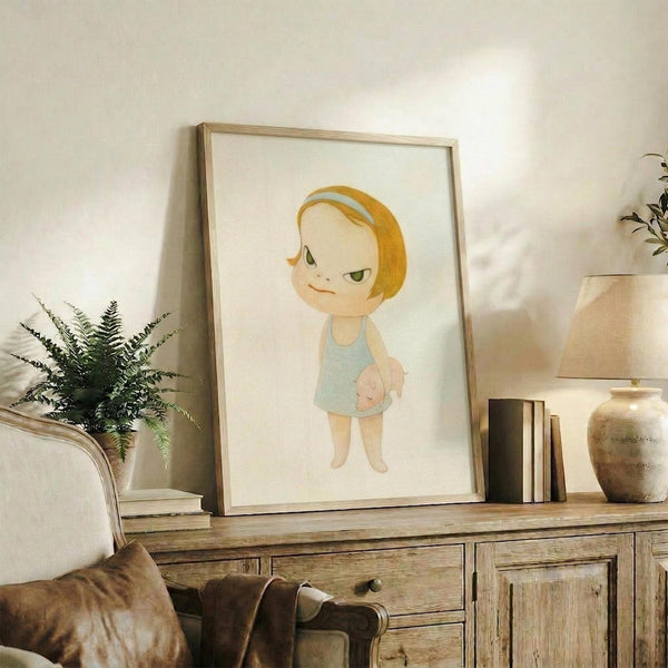 May include: Framed artwork showing a cartoon-style child character with blonde bob haircut and blue headband, wearing a light blue dress while holding a small pig. The illustration has a minimalist style with soft colors on cream background. The wooden frame sits on a rustic wood sideboard next to a fern plant, books, and ceramic vase in a neutral-toned living room setting.