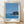 May include: Framed poster print featuring a minimalist coastal scene with blue ocean, small rocky island with dark vegetation, and clear blue sky. Modern design with bold black text below the image and five circular dots in varying shades of blue and gray. Natural wood frame with white matting, displayed on light wooden floor against white wall with soft natural lighting creating subtle shadows.