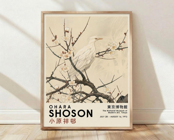 May include: Framed museum exhibition poster featuring a white egret perched on flowering plum branches against a cream background. The artwork shows delicate cherry blossoms in soft pink and white tones with dark twisted branches. Text includes "OHARA SHOSON" and Japanese characters, plus museum details. Light wood frame displayed on wooden floor against white textured wall with natural shadows.