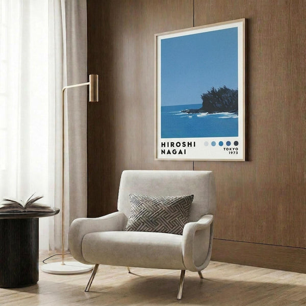 May include: A framed poster with blue ocean scene featuring a small rocky island with vegetation against clear sky, displayed on wood-paneled wall above beige upholstered armchair with geometric patterned pillow. Modern brass floor lamp and dark round side table with open book visible. Light wood frame surrounds artwork with text below ocean image showing five colored dots.