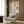 May include: Framed vintage Japanese crane poster displayed on wood-paneled wall above beige upholstered armchair with geometric pillow. White frame contains artwork showing flying crane with outstretched wings in cream and orange tones. Modern reading corner features gold floor lamp and dark round side table with open book.
