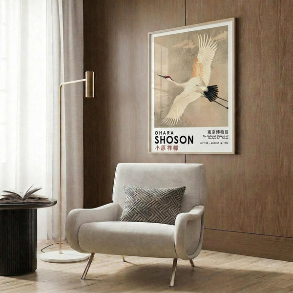 May include: Framed vintage Japanese crane poster displayed on wood-paneled wall above beige upholstered armchair with geometric pillow. White frame contains artwork showing flying crane with outstretched wings in cream and orange tones. Modern reading corner features gold floor lamp and dark round side table with open book.