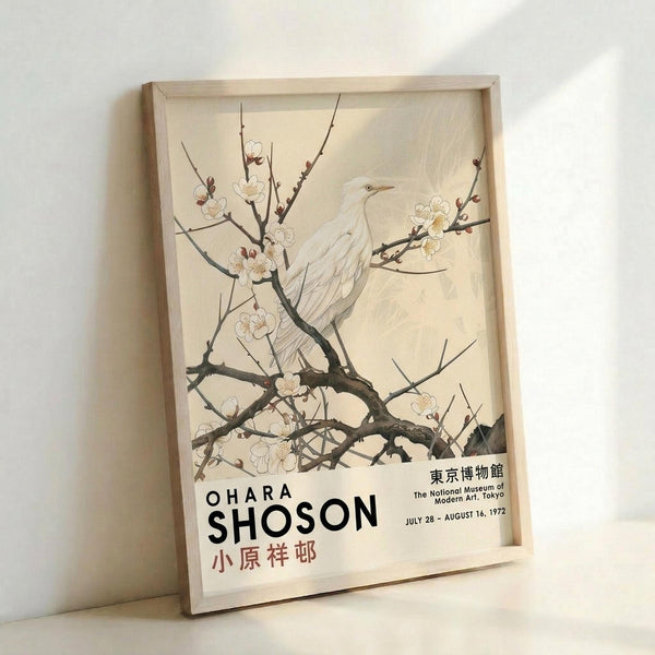 May include: Framed vintage Japanese art poster featuring a white egret perched among flowering cherry blossom branches against a cream background. Exhibition poster shows "OHARA SHOSON" text with Japanese characters and museum details. Natural wood frame leans against white wall, casting soft shadows. Traditional woodblock print style with delicate line work.
