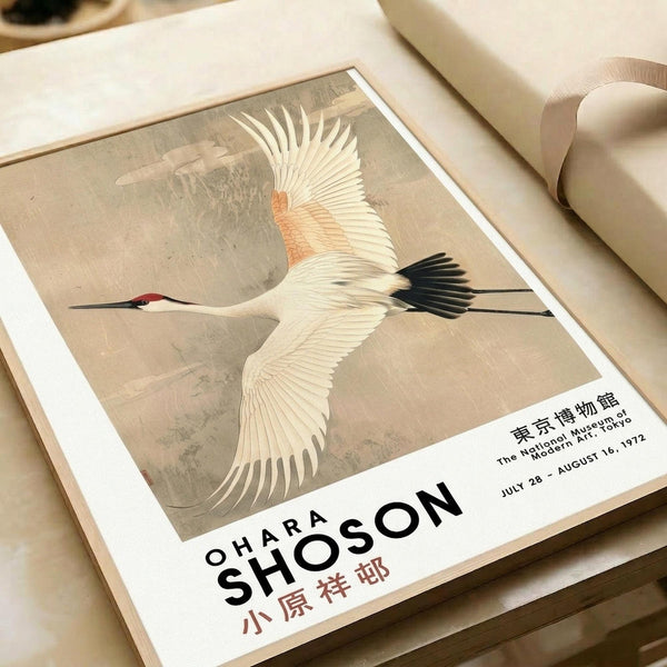 May include: Vintage-style exhibition poster featuring a white crane with black-tipped wings and red crown flying against a muted beige background. The print shows Japanese text and museum details from a 1972 Tokyo exhibition. Poster lies flat on a cream surface next to rolled paper tubes, displaying the artwork's soft earth tones and traditional Asian aesthetic.