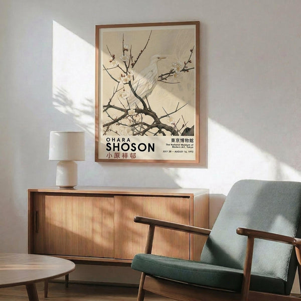 May include: Framed vintage Japanese art poster featuring white bird perched on flowering cherry blossom branches against cream background, displayed above mid-century modern wooden credenza with table lamp, next to sage green upholstered armchair with wooden arms in sunlit room with white walls