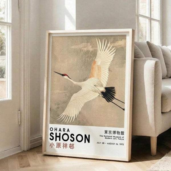 May include: A framed vintage exhibition poster featuring a white crane with outstretched wings and black wing tips against a muted beige background. The poster shows text for Ohara Shoson and The National Museum of Modern Art, Tokyo with dates July 28 - August 16, 1972. The light wood frame leans against a white wall next to a beige upholstered sofa in a bright room with large windows and wooden floors.
