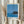 May include: Person in cream ruffled sleeves holding framed poster showing blue ocean scene with dark rocky outcrop and waves, alongside dried pampas grass stems. Print features minimalist coastal landscape in blue tones with text below reading "HIROSHI NAGAI" and "TOKYO 1973" plus color dot palette. Light wooden frame against neutral curtained background.