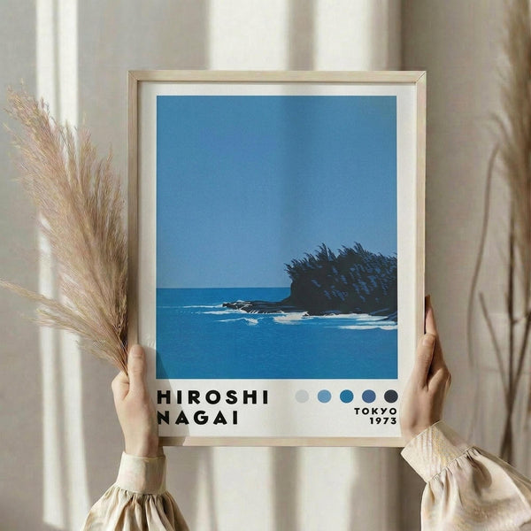 May include: Person in cream ruffled sleeves holding framed poster showing blue ocean scene with dark rocky outcrop and waves, alongside dried pampas grass stems. Print features minimalist coastal landscape in blue tones with text below reading "HIROSHI NAGAI" and "TOKYO 1973" plus color dot palette. Light wooden frame against neutral curtained background.
