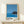 May include: Framed poster displaying a minimalist coastal scene with deep blue ocean, small rocky island with dark vegetation, and clear blue sky. Features bold black text reading "HIROSHI NAGAI" and "TOKYO 1973" with four circular color dots. Natural wood frame with white matting, positioned on light wooden shelf alongside small wrapped gift with twine and evergreen sprig against cream wall.