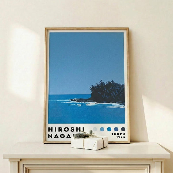 May include: Framed poster displaying a minimalist coastal scene with deep blue ocean, small rocky island with dark vegetation, and clear blue sky. Features bold black text reading "HIROSHI NAGAI" and "TOKYO 1973" with four circular color dots. Natural wood frame with white matting, positioned on light wooden shelf alongside small wrapped gift with twine and evergreen sprig against cream wall.