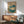 May include: Vintage Japanese botanical artwork showing lotus flowers with cream petals and large teal-blue leaves, displayed in wooden frame above mid-century modern credenza. Sage green upholstered chair with wood arms and round coffee table visible in foreground. Warm sunlight creates shadows on white wall, with ceramic table lamp on sideboard.