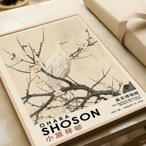 May include: Vintage Japanese art poster featuring a white heron perched on flowering plum tree branches with delicate white blossoms, displayed in warm cream matting alongside rolled paper tubes on a neutral surface. The artwork shows traditional Asian brushwork style with subtle earth tones and includes museum exhibition text at bottom.