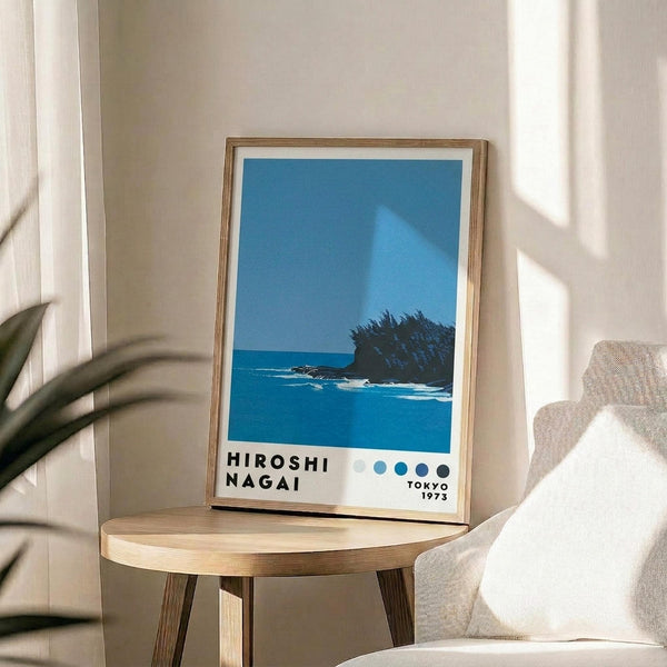 May include: Framed poster print displaying a minimalist seascape with bright blue water and small rocky island with dark vegetation under clear sky. Text shows "HIROSHI NAGAI" and "TOKYO 1973" with four small dots. Thin natural wood frame with white mat, positioned on round wooden side table next to beige upholstered chair. Soft natural lighting creates shadows on cream wall with plant leaves visible in foreground.