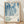 May include: Framed poster leaning against white wall on wooden floor. Artwork features abstract blue and white organic shapes with dotted patterns and circular forms. Text below reads "YAYOI KUSAMA" with Japanese characters, "Tokyo, Japan 1984" and museum name. Light wooden frame surrounds cream-colored print. Natural lighting creates soft shadows on textured white wall.