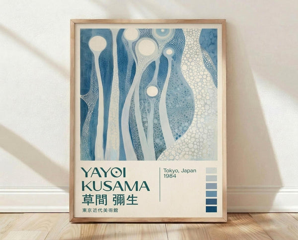 May include: Framed poster leaning against white wall on wooden floor. Artwork features abstract blue and white organic shapes with dotted patterns and circular forms. Text below reads "YAYOI KUSAMA" with Japanese characters, "Tokyo, Japan 1984" and museum name. Light wooden frame surrounds cream-colored print. Natural lighting creates soft shadows on textured white wall.