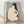 May include: Framed Japanese woodblock print depicting a nude woman with long black hair combing her hair, rendered in soft peach and blue-green tones with traditional calligraphy and red stamps, displayed in light wooden frame leaning against white wall on wooden floor with natural lighting casting shadows