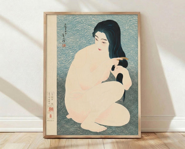 May include: Framed Japanese woodblock print depicting a nude woman with long black hair combing her hair, rendered in soft peach and blue-green tones with traditional calligraphy and red stamps, displayed in light wooden frame leaning against white wall on wooden floor with natural lighting casting shadows