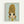 Framed artwork showing a cartoon-style character with an exaggerated tall golden blonde beehive hairdo and large dark eyes against a pale mint green background. The figure has a melancholic expression with bangs covering part of their face and wears a yellow-green top. The piece is displayed in a simple light wood frame on a white wall, rendered in a distinctive Japanese pop art aesthetic with muted earth tones.
