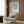 May include: Framed Yayoi Kusama exhibition poster with blue abstract organic shapes and Japanese text, displayed on wood-paneled wall above beige upholstered armchair with geometric throw pillow. Modern brass floor lamp and dark round side table with open book complete the contemporary living room setting with warm lighting.