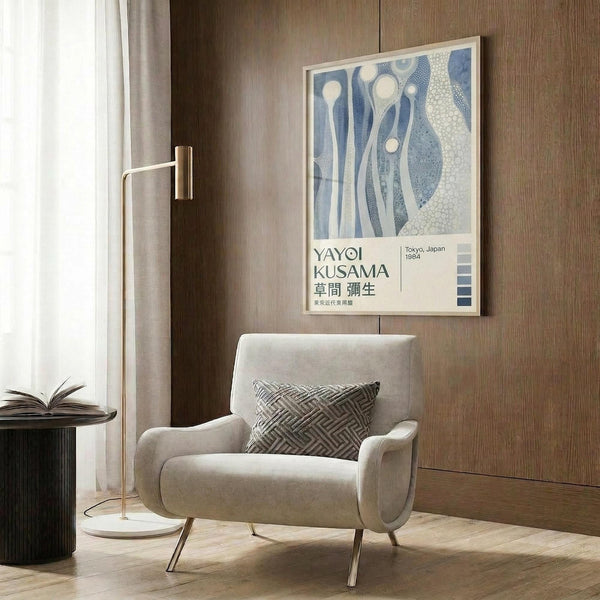 May include: Framed Yayoi Kusama exhibition poster with blue abstract organic shapes and Japanese text, displayed on wood-paneled wall above beige upholstered armchair with geometric throw pillow. Modern brass floor lamp and dark round side table with open book complete the contemporary living room setting with warm lighting.