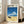 May include: Framed poster print leaning against beige upholstered chair in bright living room with white walls and windows. Artwork shows vintage black sedan on sandy beach with blue ocean waves and puffy white clouds against deep blue sky. Poster has white border with black text at top including Japanese characters. Natural wood frame, approximately 2-3 feet tall, casting soft shadows on hardwood floor.