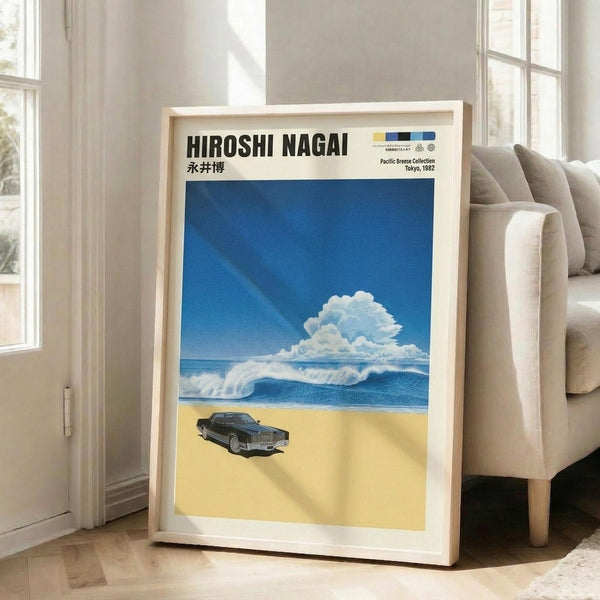 May include: Framed poster print leaning against beige upholstered chair in bright living room with white walls and windows. Artwork shows vintage black sedan on sandy beach with blue ocean waves and puffy white clouds against deep blue sky. Poster has white border with black text at top including Japanese characters. Natural wood frame, approximately 2-3 feet tall, casting soft shadows on hardwood floor.