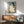 May include: Japanese woodblock print showing woman with long black hair combing, seated figure in cream tones against teal background, displayed in thin wood frame above mid-century modern walnut credenza with ceramic table lamp, next to gray upholstered armchair with wooden arms, white walls with dramatic window light shadows