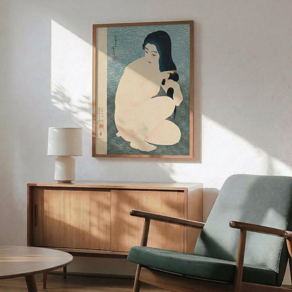 May include: Japanese woodblock print showing woman with long black hair combing, seated figure in cream tones against teal background, displayed in thin wood frame above mid-century modern walnut credenza with ceramic table lamp, next to gray upholstered armchair with wooden arms, white walls with dramatic window light shadows