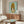 May include: Framed poster of anime-style character with blonde beehive hair and large eyes on mint green background, displayed above wooden sideboard in modern living room with white curved chair, copper floor lamp, and glass coffee table