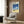 May include: A framed vintage-style poster featuring a dark car on a golden beach with blue ocean waves and white clouds against a clear sky. The poster displays "HIROSHI NAGAI" text at the top. Shown hanging on wood-paneled wall above a cream upholstered armchair with geometric patterned pillow, beside a brass floor lamp and dark round side table with open book.