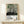 May include: Framed art print leaning against white wall on light wood shelf, featuring abstract forest scene with black tree trunks, sage green circular shapes, and dotted ground in cream, green and black. Small wrapped gift box sits in front. Thin wood frame, modern minimalist setting with natural lighting.