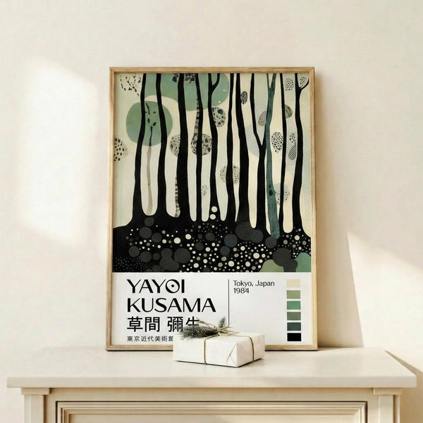 May include: Framed art print leaning against white wall on light wood shelf, featuring abstract forest scene with black tree trunks, sage green circular shapes, and dotted ground in cream, green and black. Small wrapped gift box sits in front. Thin wood frame, modern minimalist setting with natural lighting.
