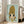 May include: Large framed artwork leaning against beige sofa in bright living room with white walls and windows. Art shows anime-style character with oversized blonde beehive hairstyle, large dark eyes, and yellow dress on mint green background. Light wood frame, displayed on hardwood floors with natural lighting.
