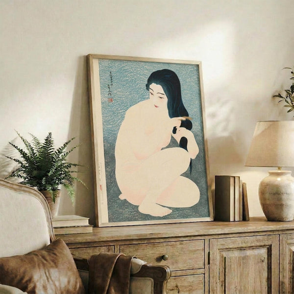 May include: Japanese woodblock print style artwork in light wood frame showing nude woman with long black hair combing her hair against turquoise background. Print displayed on rustic wooden sideboard with books, ceramic vases, fern plant, and leather armchair visible in warm-toned living room setting.